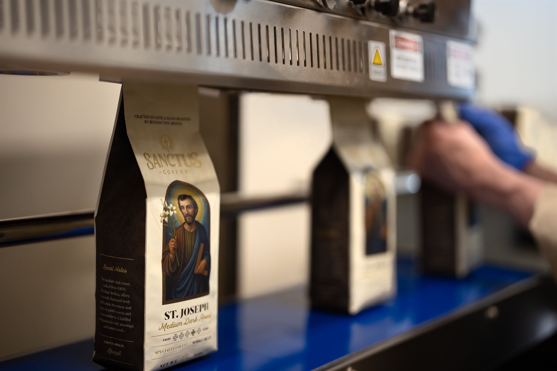 St. Joseph Sanctus Coffee bags on the production packaging line with Benedictine monk working in the background